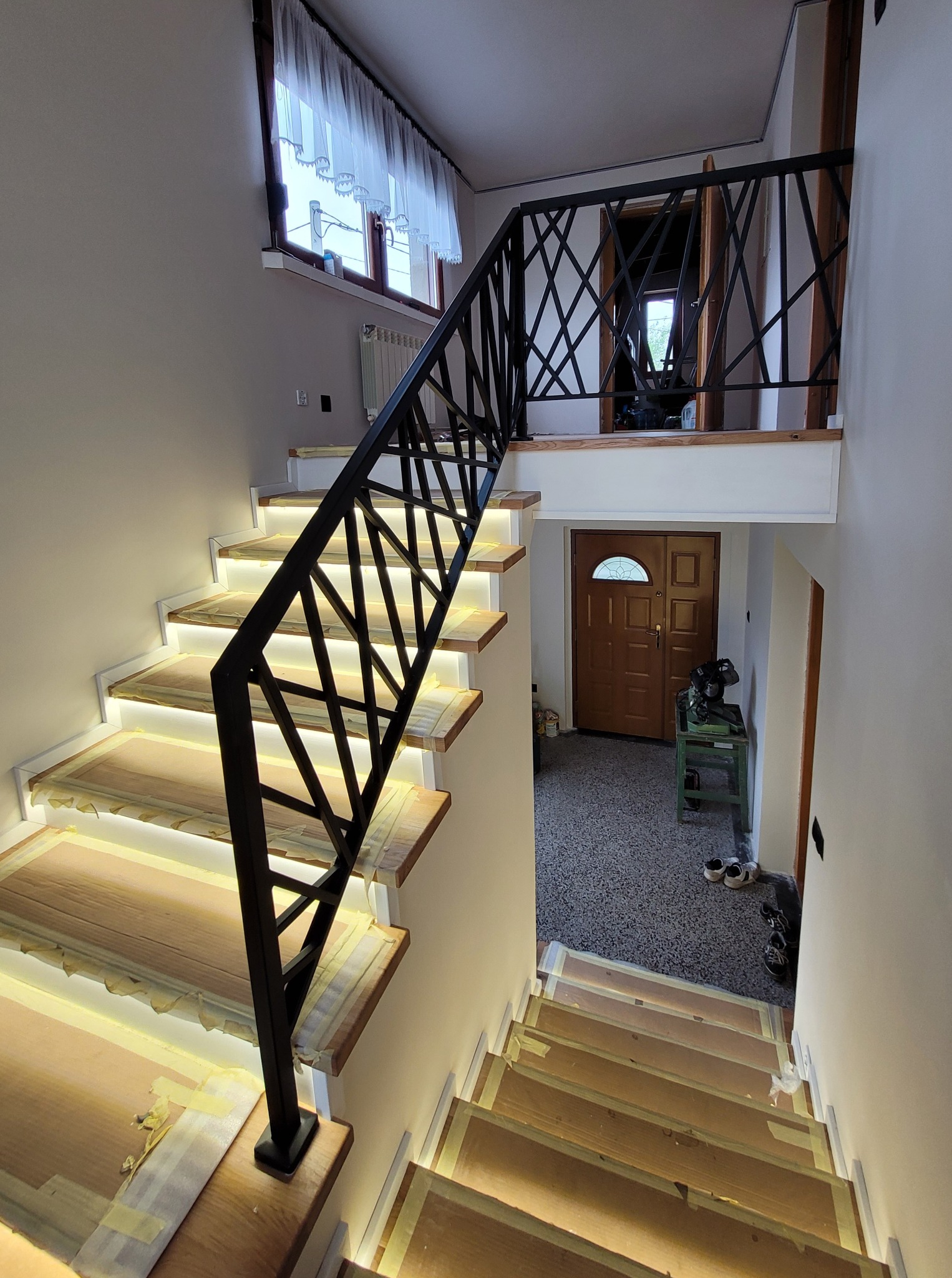 Wnętrze domu z drewnianymi schodami podświetlonymi taśmą LED i czarną metalową balustradą o geometrycznym wzorze, widok z góry na dół, etap wykończeniowy zabezpieczony taśmą malarską.