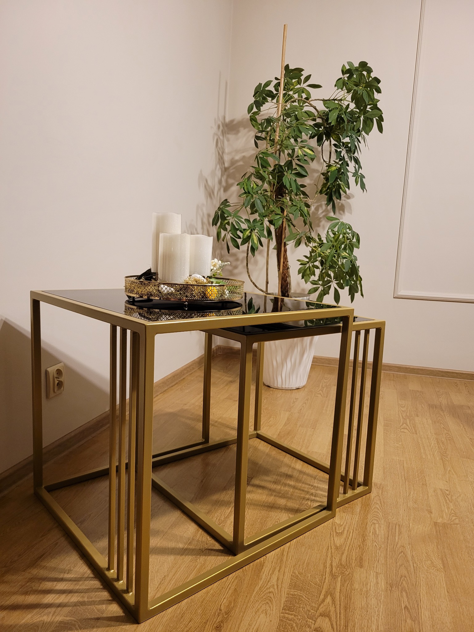 Dwa złote, metalowe stoliki kawowe w stylu loft z czarnymi blatami, ustawione jeden pod drugim. Na większym stoliku dekoracyjny złoty talerz z białymi świecami i drobnymi ozdobami. W tle roślina...