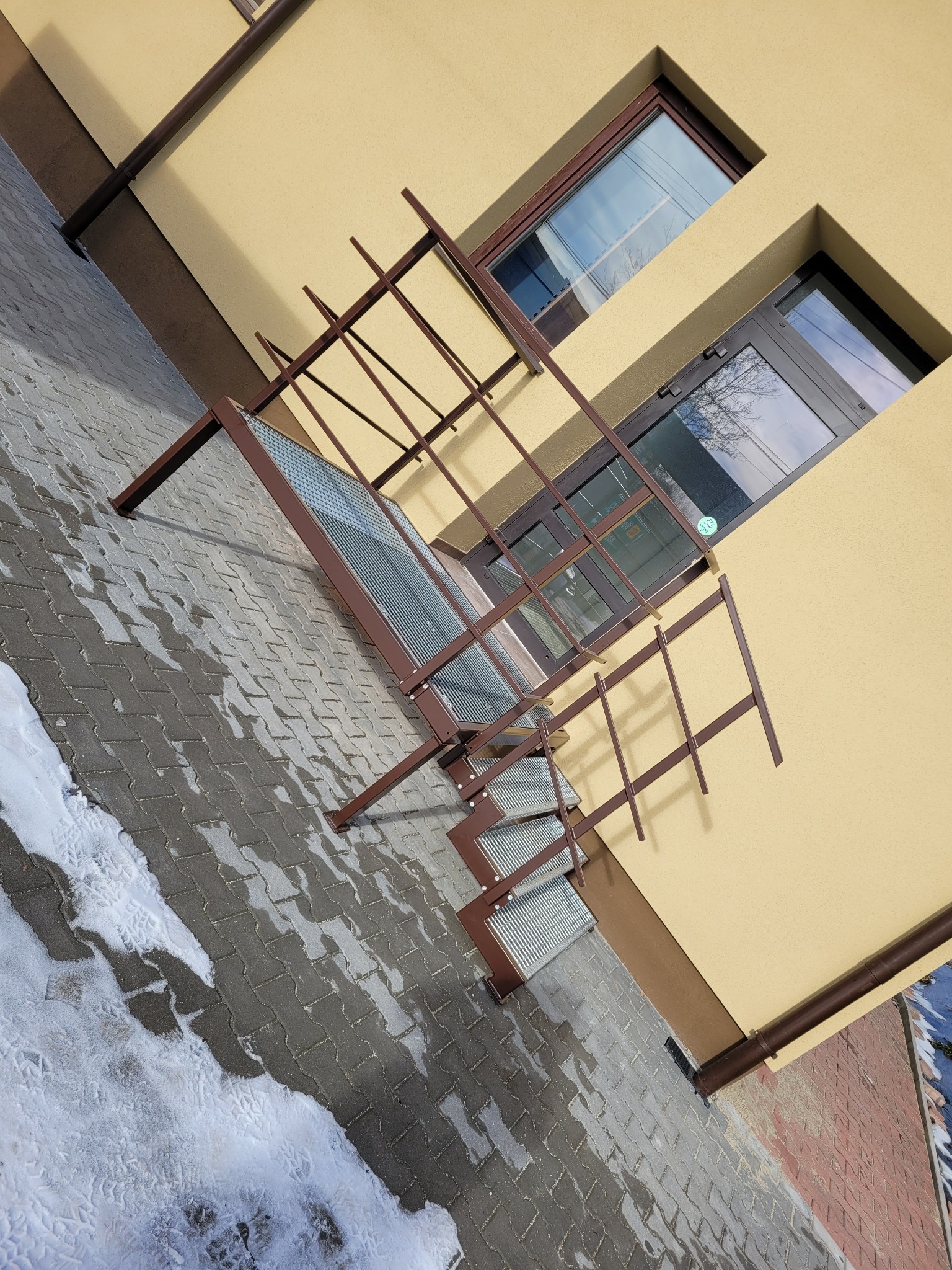 Zewnętrzne metalowe schody przeciwpożarowe z kratownicowymi stopniami, przymocowane do elewacji budynku w pobliżu okien, z widocznym śniegiem na brukowanej nawierzchni.