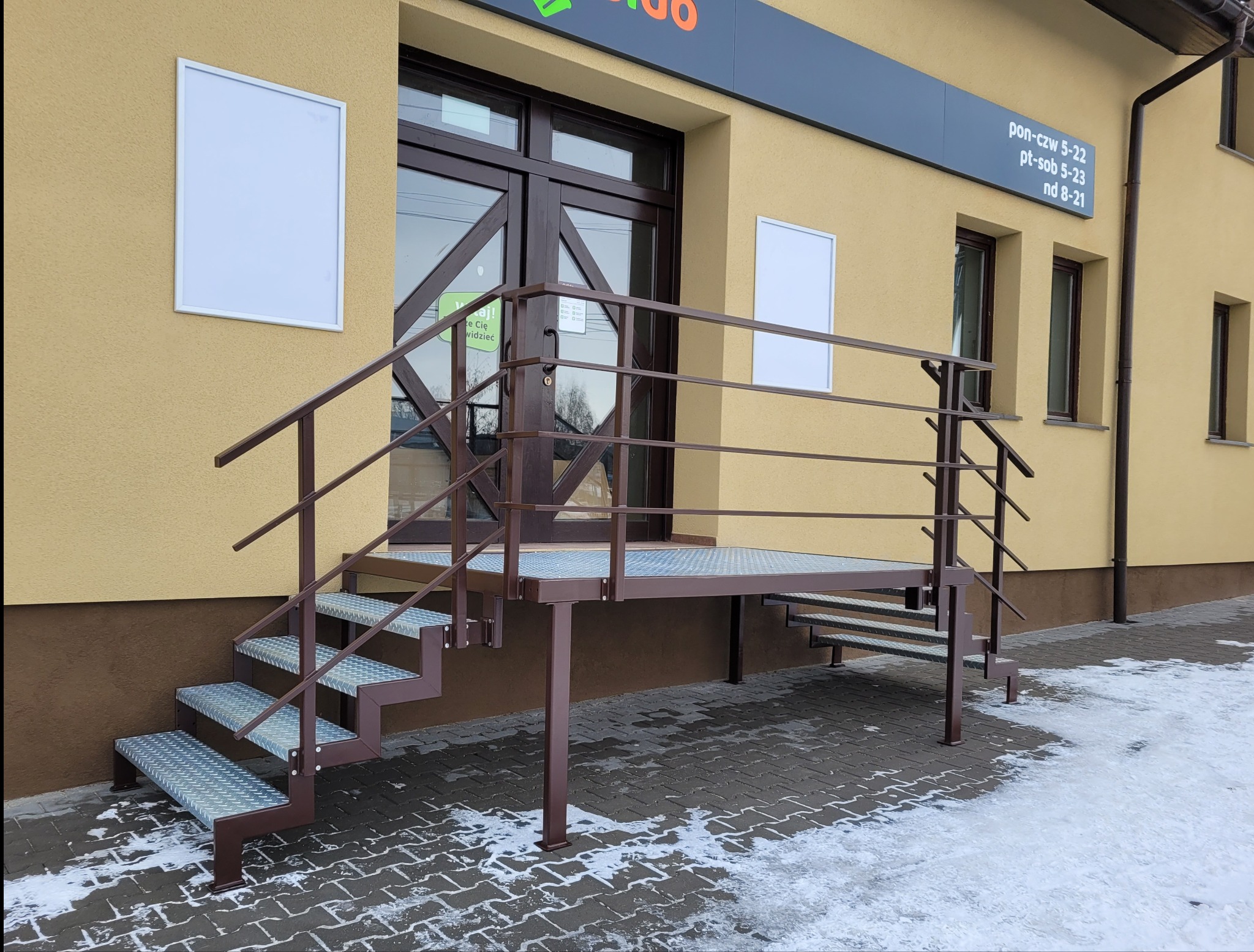 Zewnętrzne metalowe schody z podestem prowadzące do wejścia budynku, brązowa konstrukcja, antypoślizgowe stopnie, częściowo ośnieżony chodnik.