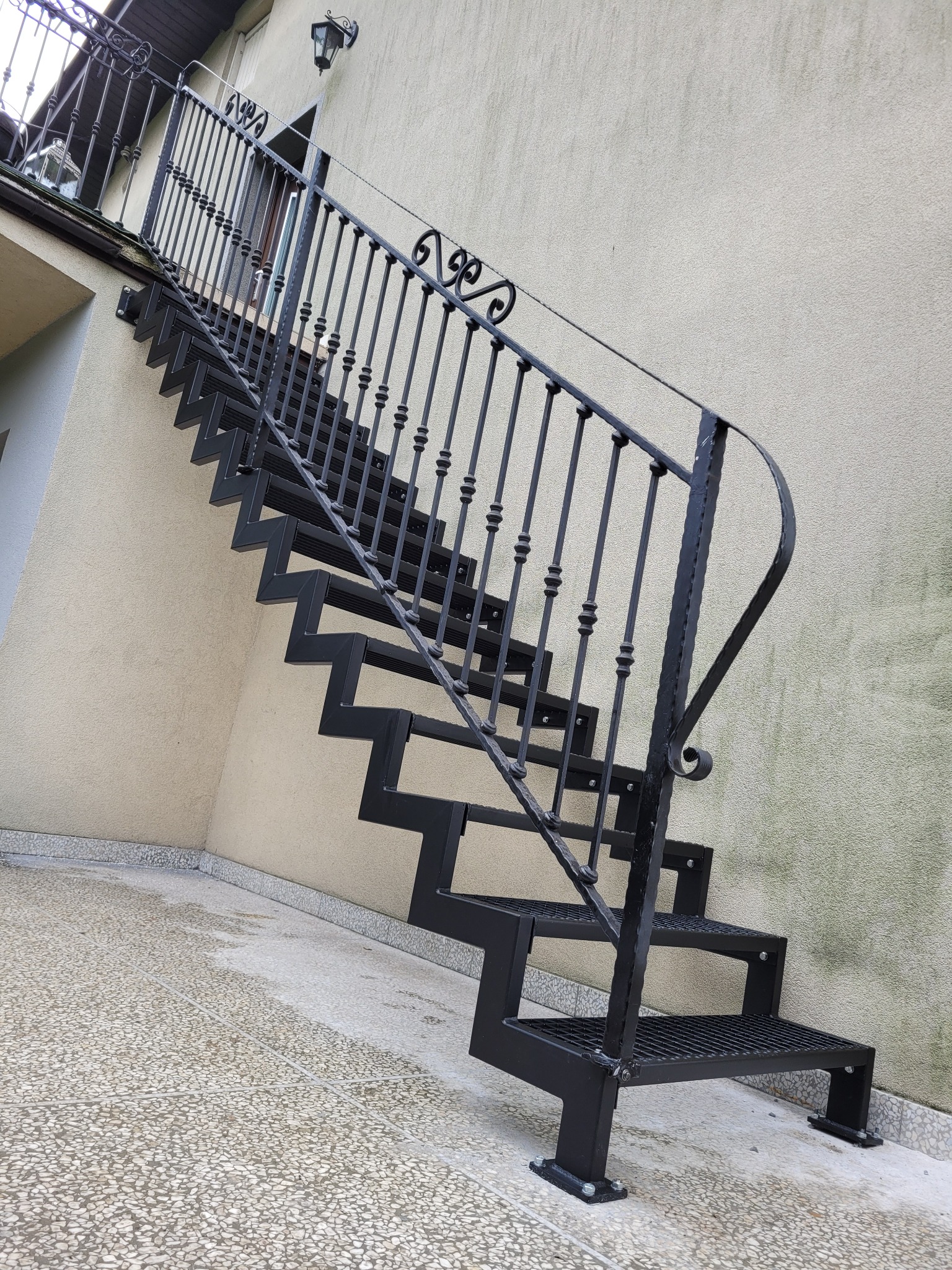 Zewnętrzne schody metalowe z czarną balustradą, prowadzące do balkonu na tle beżowej ściany budynku.