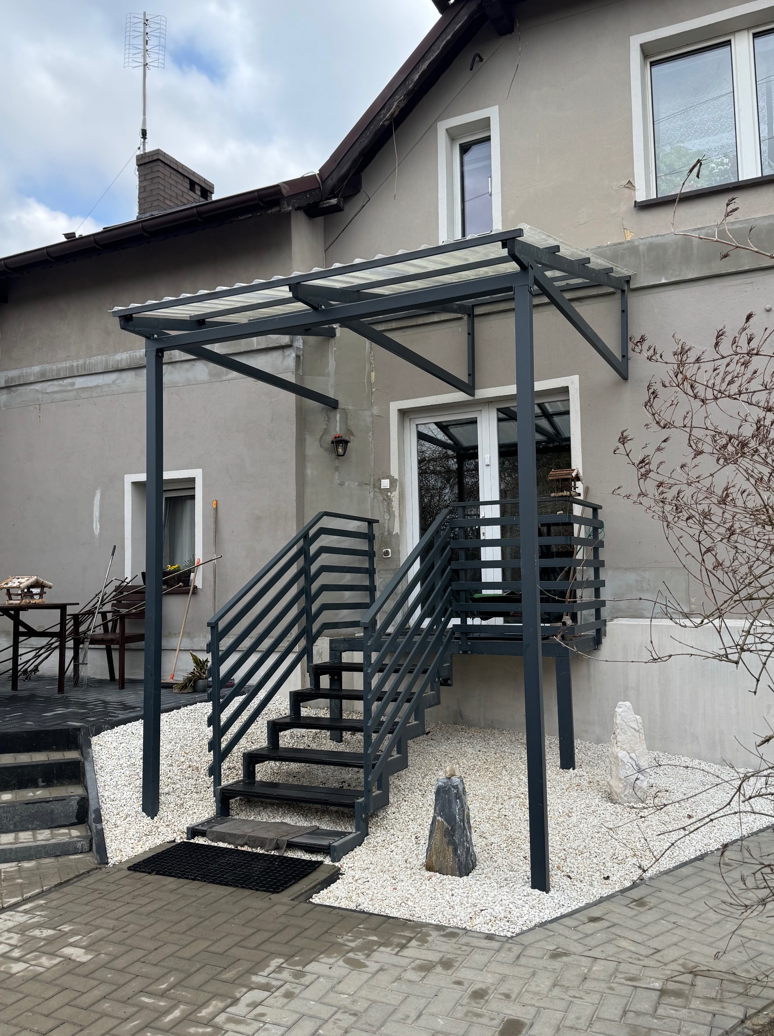 Metalowe schody zewnętrzne z zadaszeniem z poliwęglanu prowadzące do drzwi. Białe kamienie dekoracyjne u podnóża schodów. Szara elewacja budynku.