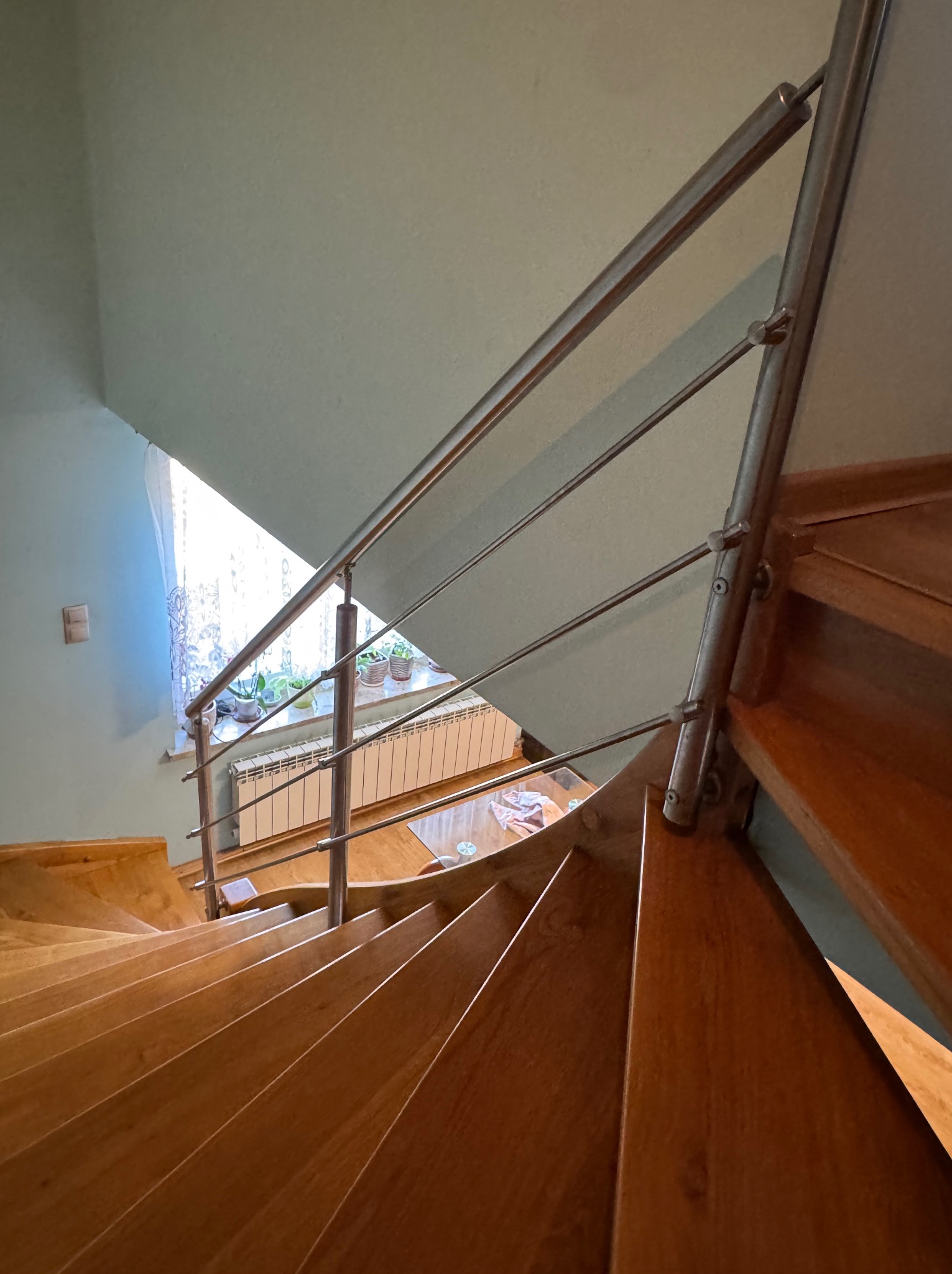 Drewniane schody spiralne z metalową balustradą, widok z góry. Na parapecie okna widoczne rośliny w doniczkach. Schody w odcieniach brązu, ściany jasne.