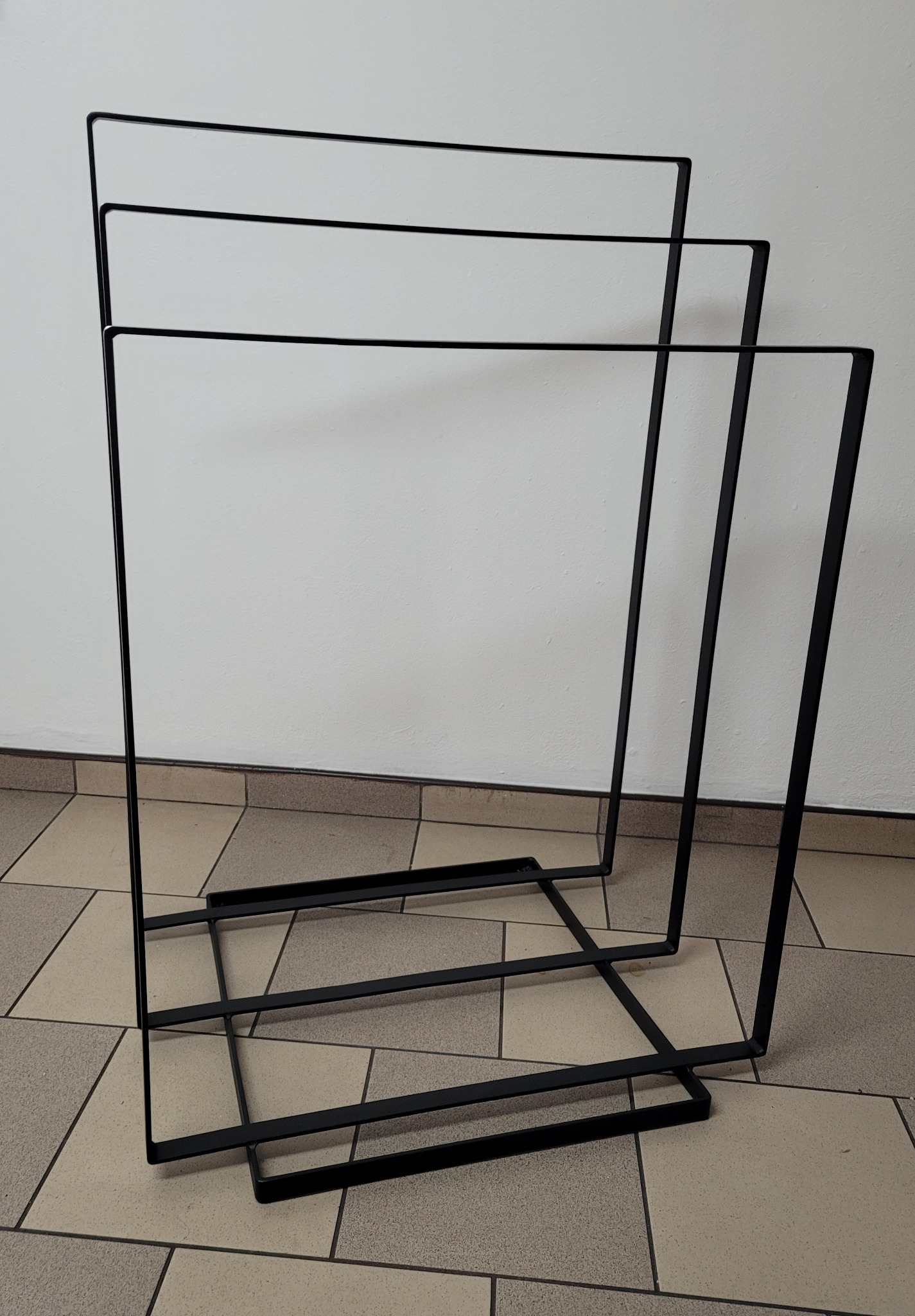 Czarna, metalowa konstrukcja przypominająca stojak na ręczniki lub parawan, składająca się z prostokątnych ram, ustawiona na kafelkach pod ścianą.