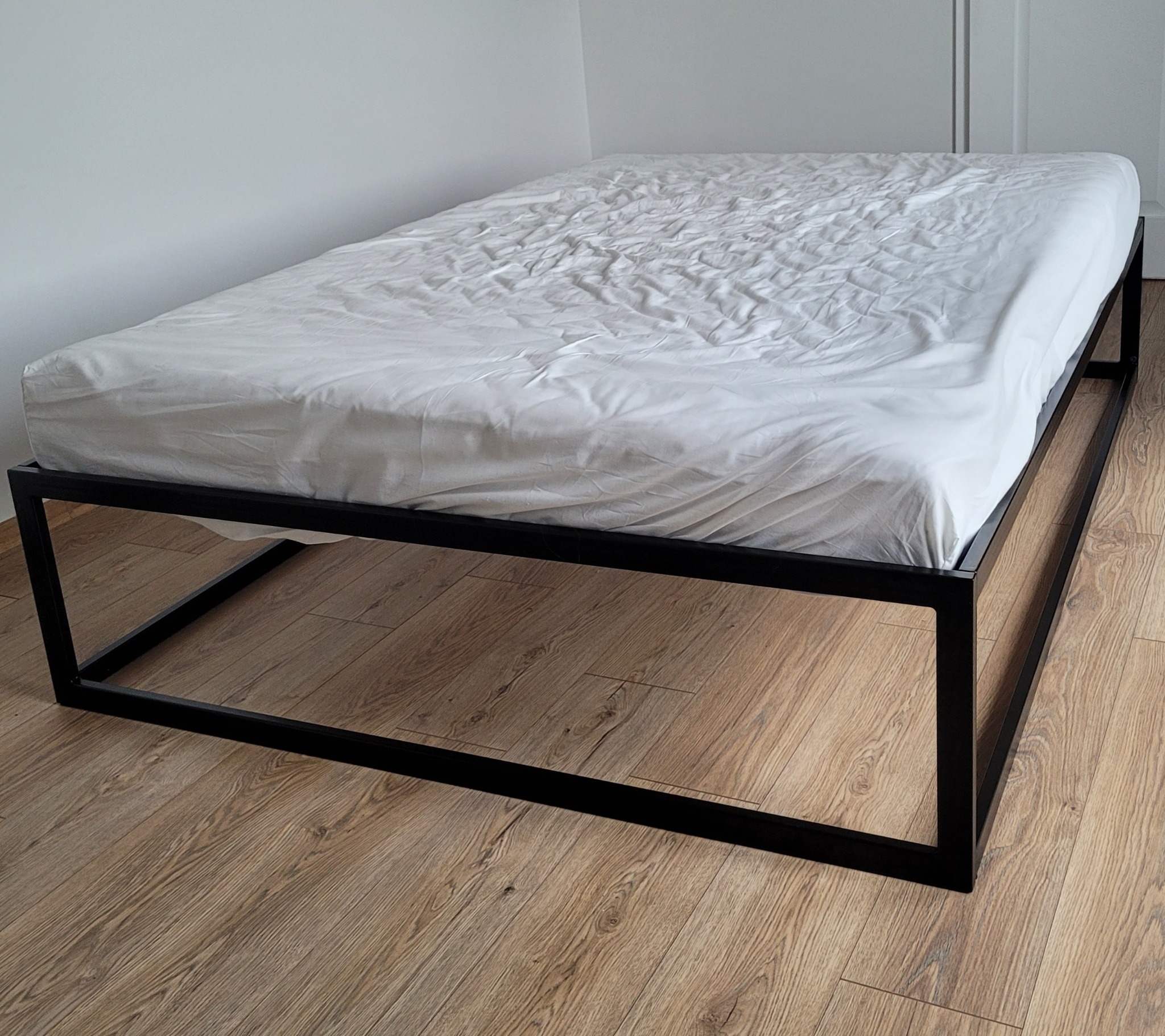 Minimalistyczna rama łóżka metalowego z czarnych profili kwadratowych, przykryta białą, pogniecioną pościelą, na podłodze z paneli drewnopodobnych.