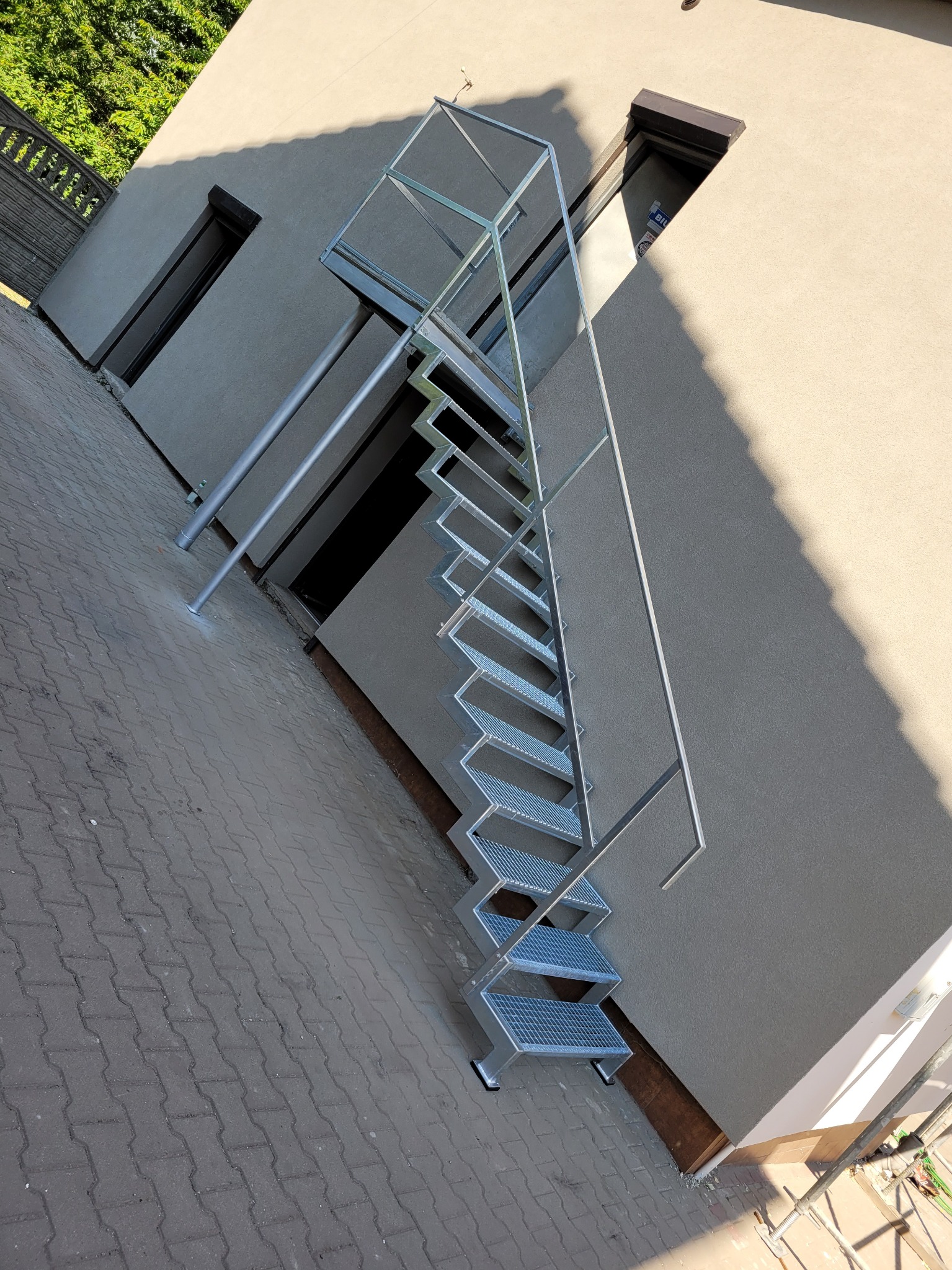 Zewnętrzne metalowe schody kratowe prowadzące do piwnicy w budynku o jasnej elewacji, widoczne z poziomu kostki brukowej.