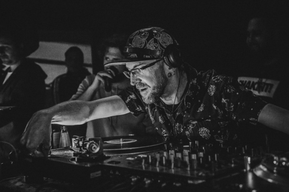 Czarno-białe zdjęcie DJ-a w czapce z wzorem paisley i słuchawkach, miksującego muzykę na gramofonie w ciemnym klubie, z widocznymi sylwetkami publiczności w tle.