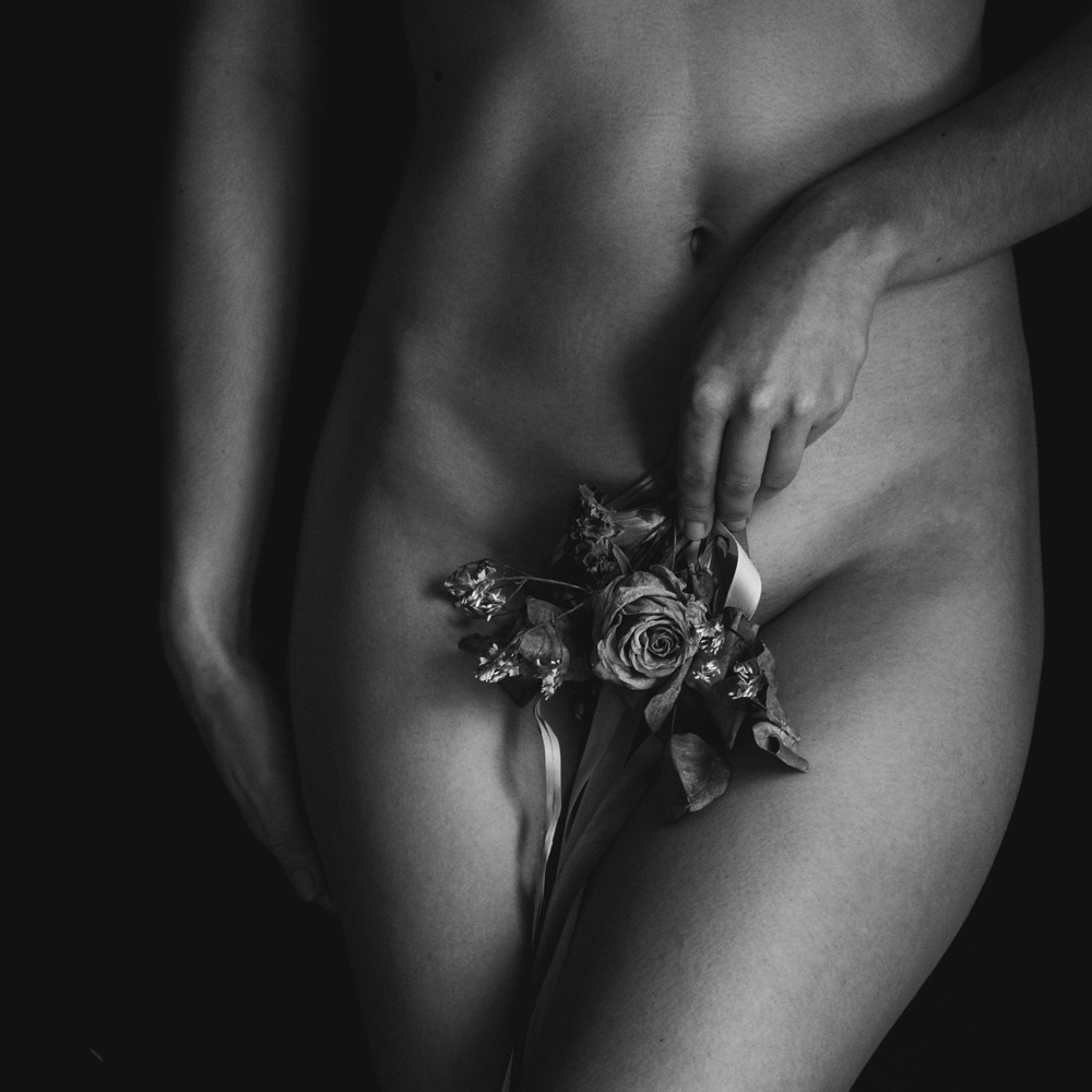 Czarno-białe zbliżenie nagiego ciała kobiety z więdnącymi kwiatami róży i wstążką, trzymanymi w dłoniach, na ciemnym tle.