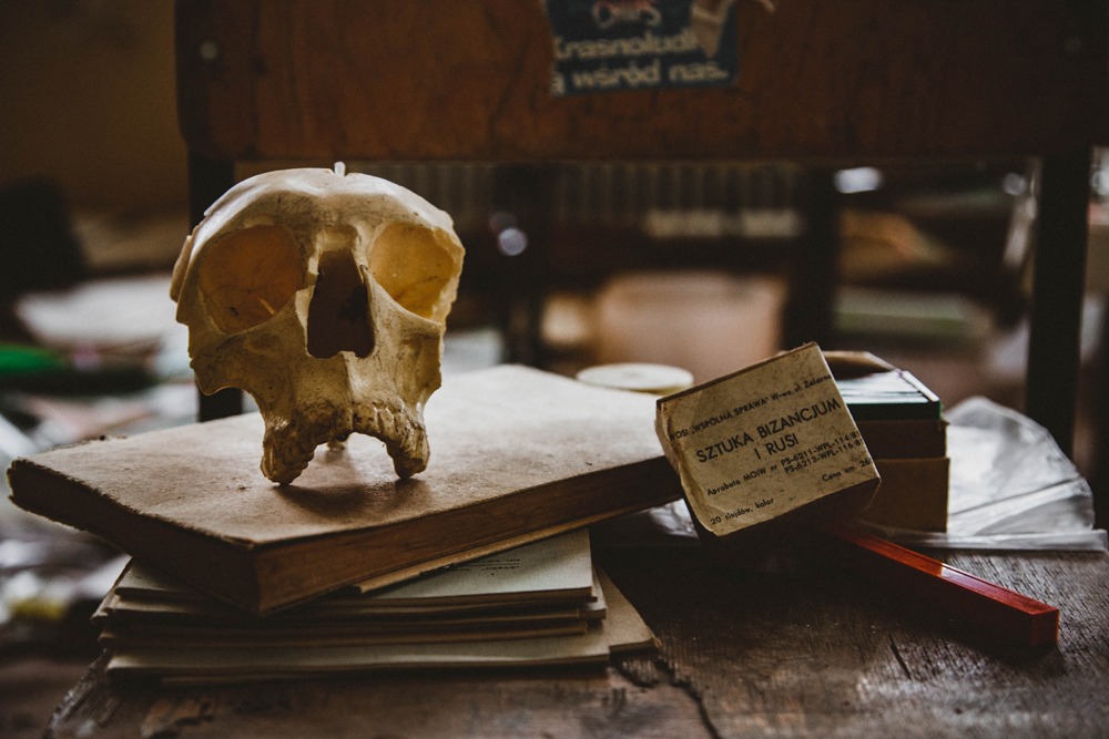 Stylizowana czaszka na stercie starych książek i pudełko z napisem 'Sztuka Bizancjum i Rusi' na zniszczonym drewnianym stole, w tle krzesło i napis na ścianie.