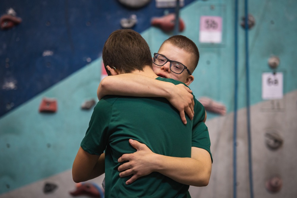 Dwóch mężczyzn w zielonych koszulkach obejmuje się na tle ściany wspinaczkowej z kolorowymi chwytami.