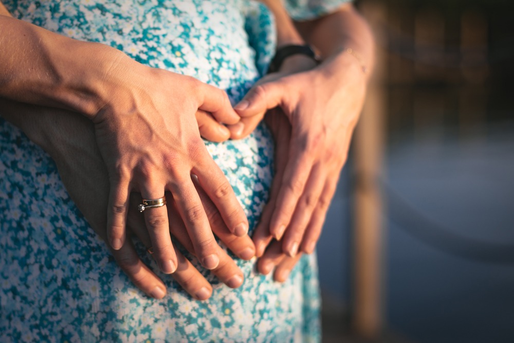 Brzuch kobiety w kwiecistej sukience, dłonie partnera i partnerki ułożone w kształt serca, pierścionek zaręczynowy na palcu.