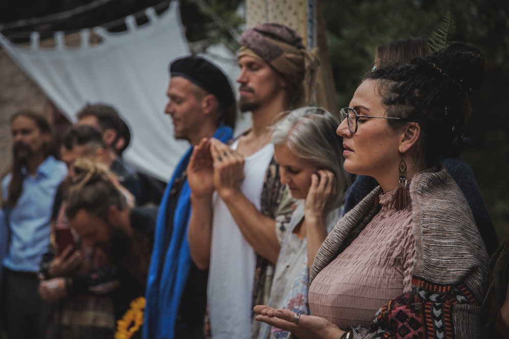 Grupa osób w różnym wieku, ubrana w stylu boho, stoi w skupieniu z zamkniętymi oczami, prawdopodobnie podczas ceremonii na zewnątrz, z białym materiałem i zielenią w tle.