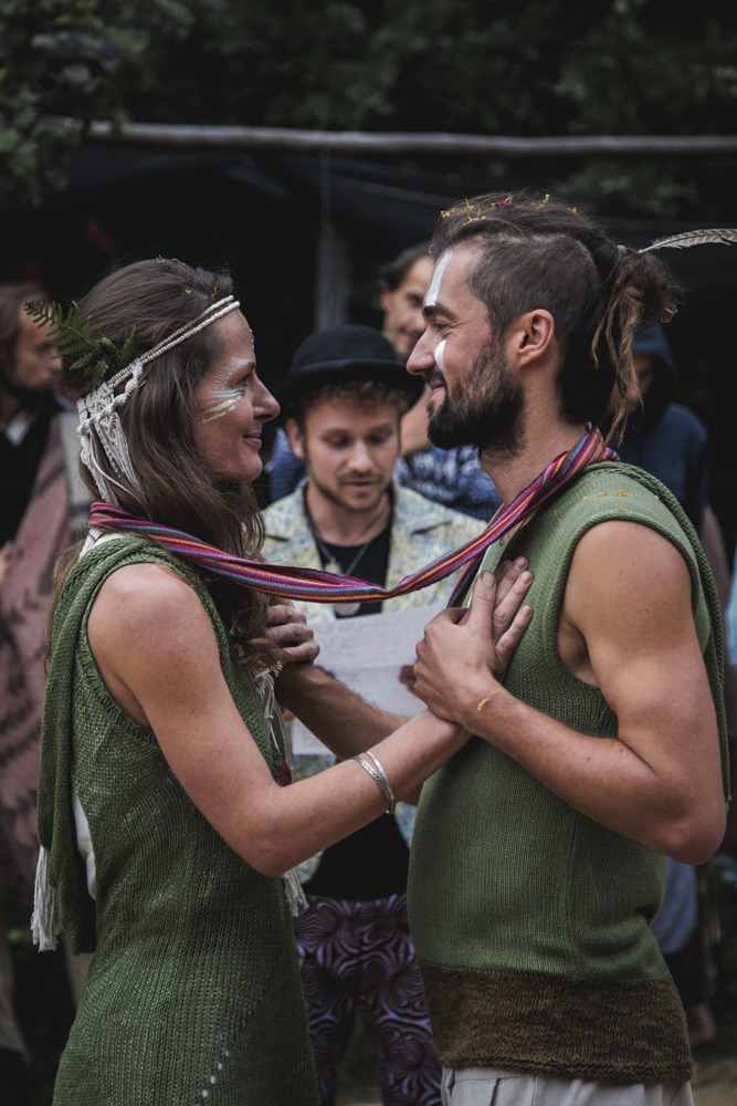 Para młoda w stylu rustykalnym, z twarzami pomalowanymi białymi paskami, trzymająca się za ręce podczas ceremonii ślubnej na łonie natury, z gośćmi w tle.