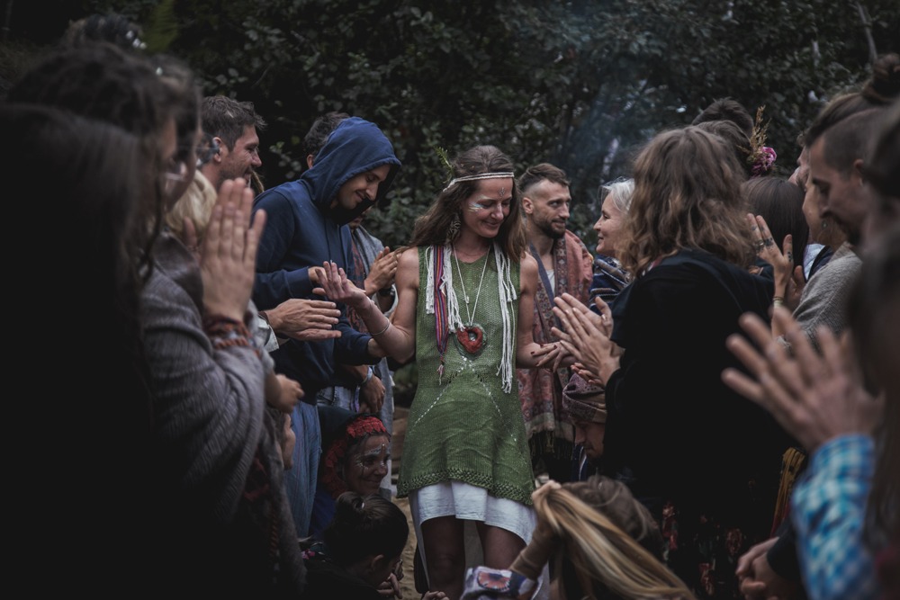 Kobieta w zielonej sukience otoczona przez ludzi klaszczących w dłonie na tle zieleni, z elementami dekoracji na twarzy i w włosach, w otoczeniu uśmiechniętych osób.