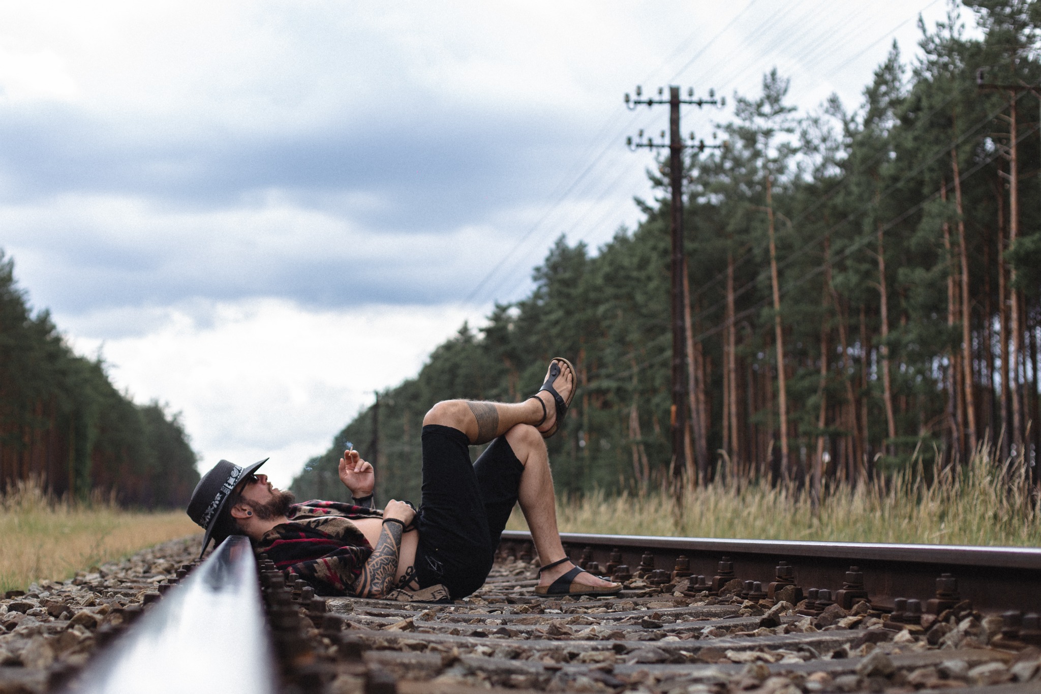 Mężczyzna z tatuażami, w kapeluszu i koszuli w kratę, leży na torach kolejowych i pali papierosa, w tle las i pochmurne niebo.