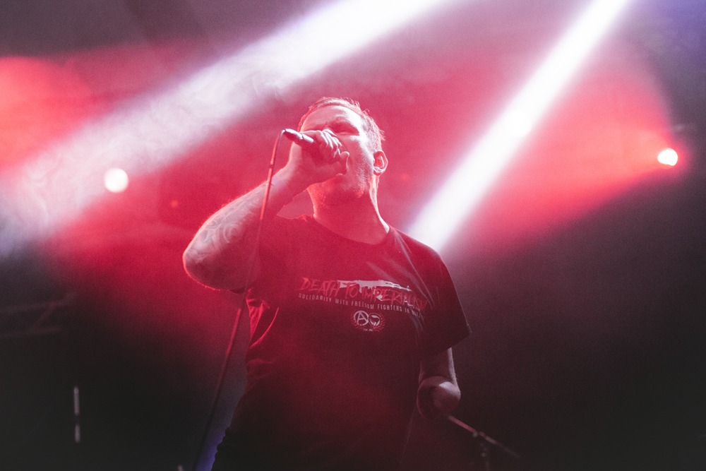 Wokalista z tatuażem na ramieniu śpiewa do mikrofonu na scenie oświetlonej czerwonymi i białymi reflektorami.