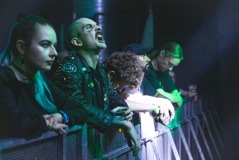 Widzowie podczas koncertu: kobieta z niebieskimi włosami, mężczyzna z wytatuowaną twarzą i skórzaną kurtką z ćwiekami, oraz inni fani opierający się o barierkę pod zielonym oświetleniem scenicznym.