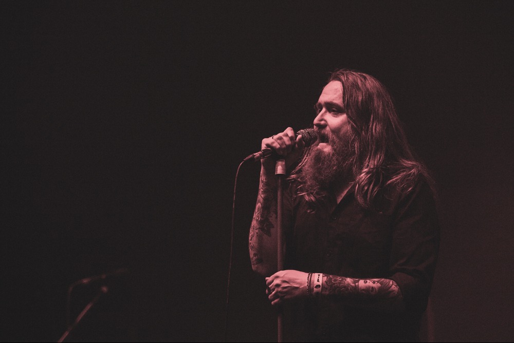 Mężczyzna z brodą i tatuażami na ramionach śpiewa do mikrofonu na scenie w ciemnym, czerwonawym oświetleniu.