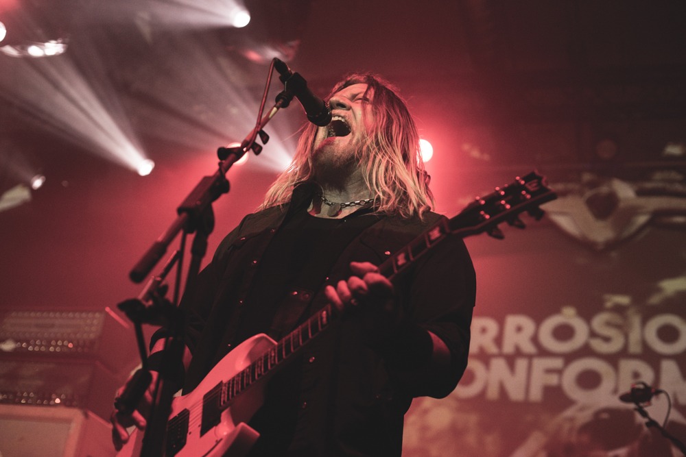 Wokalista z długimi blond włosami, w czarnym stroju, śpiewa do mikrofonu i gra na białej gitarze elektrycznej na scenie oświetlonej czerwonymi reflektorami.