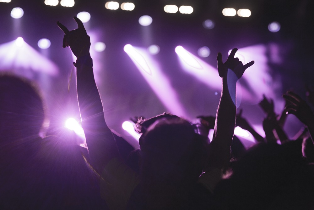 Tłum ludzi na koncercie, wzniesione ręce w geście rocka, fioletowe światła sceniczne tworzą dynamiczną atmosferę.