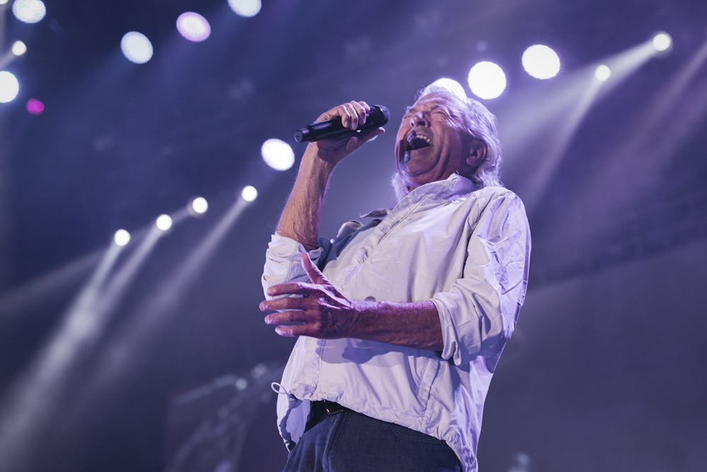 Starszy mężczyzna z mikrofonem śpiewa na scenie w jasnym, fioletowym oświetleniu, z uniesioną ręką i szeroko otwartymi ustami.
