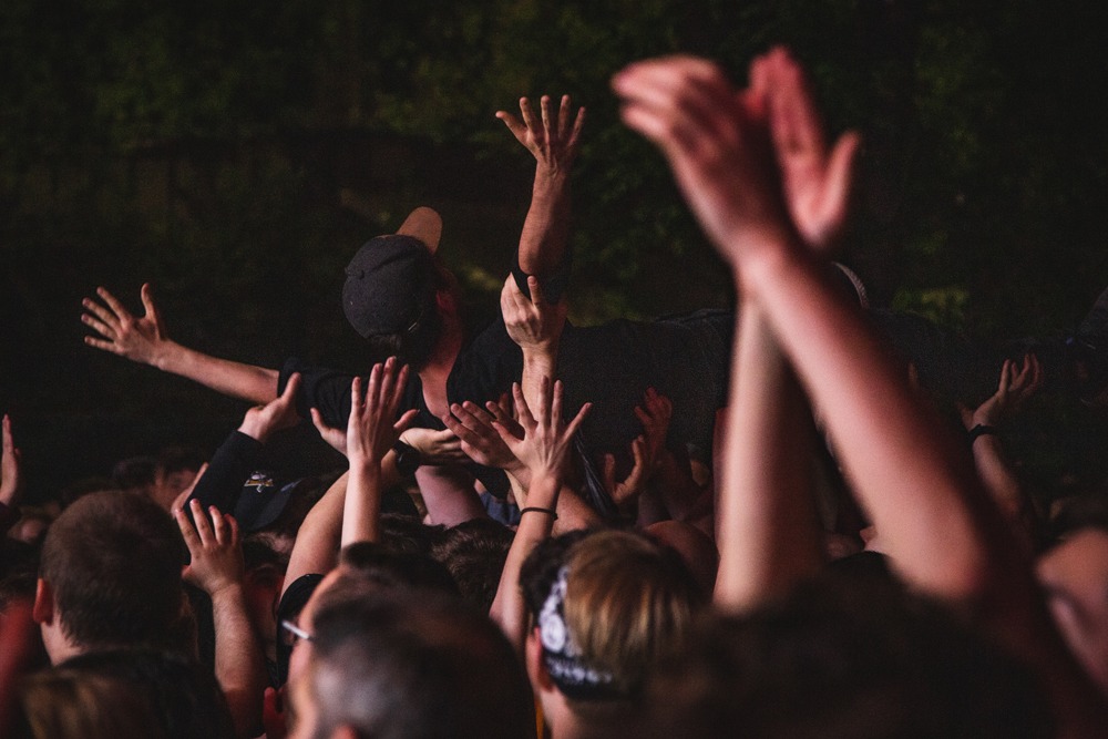 Tłum fanów podnosi na rękach muzyka podczas koncertu, widoczne liczne dłonie i fragmenty twarzy publiczności, ciemne tło.