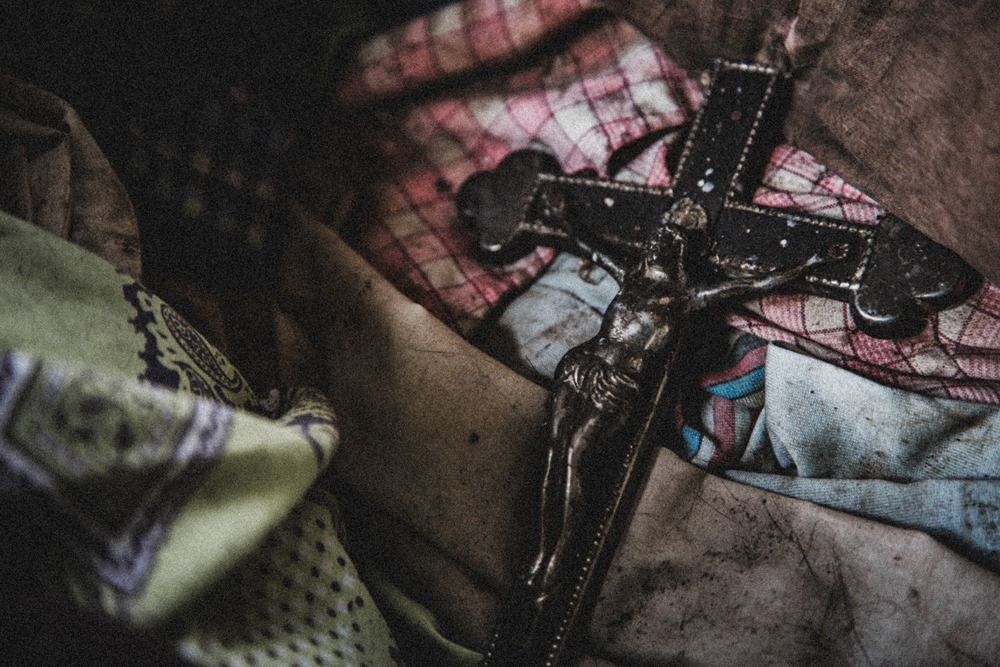 Metalowy krucyfiks z Jezusem na tle pogniecionej, wzorzystej tkaniny w odcieniach czerwieni, błękitu i zieleni, w ciemnym otoczeniu.