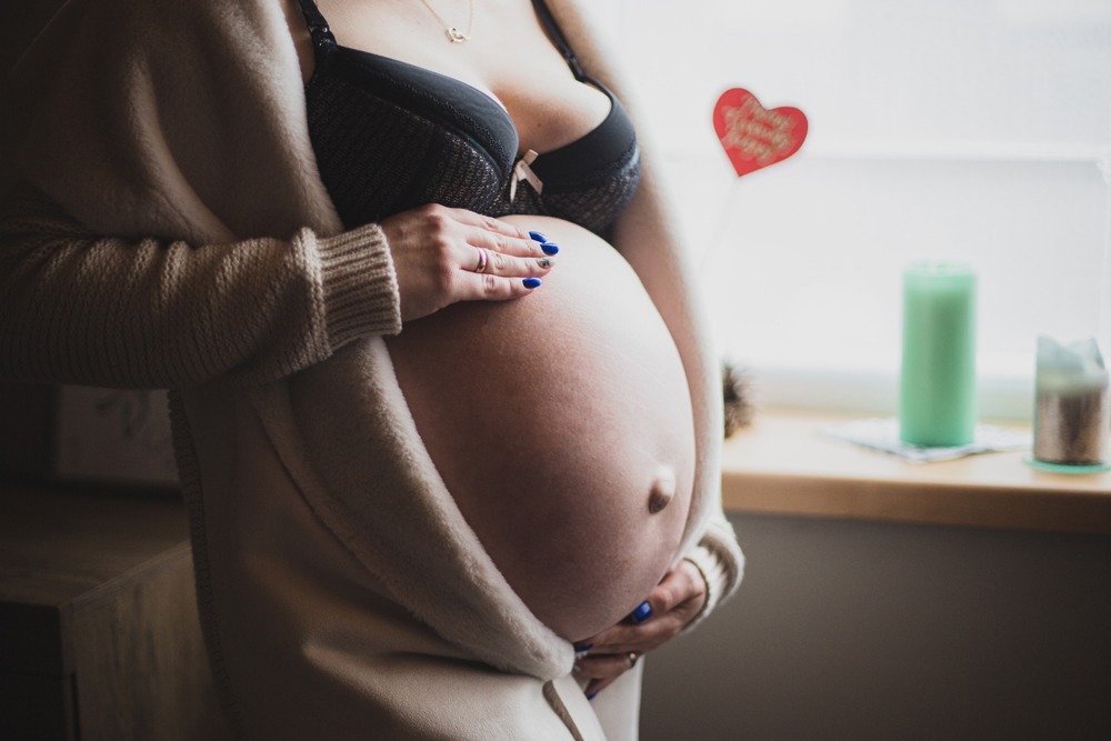 Kobieta w czarnym biustonoszu i beżowym swetrze trzyma ręce na dużym brzuchu ciążowym, z błękitnym manicure, w tle okno z zieloną świecą i dekoracją w kształcie serca.