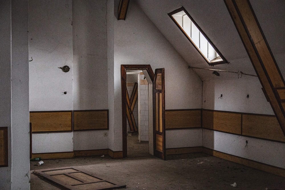 Opuszczone poddasze z otwartymi drzwiami, widoczne fragmenty boazerii, okno dachowe i resztki instalacji elektrycznej.