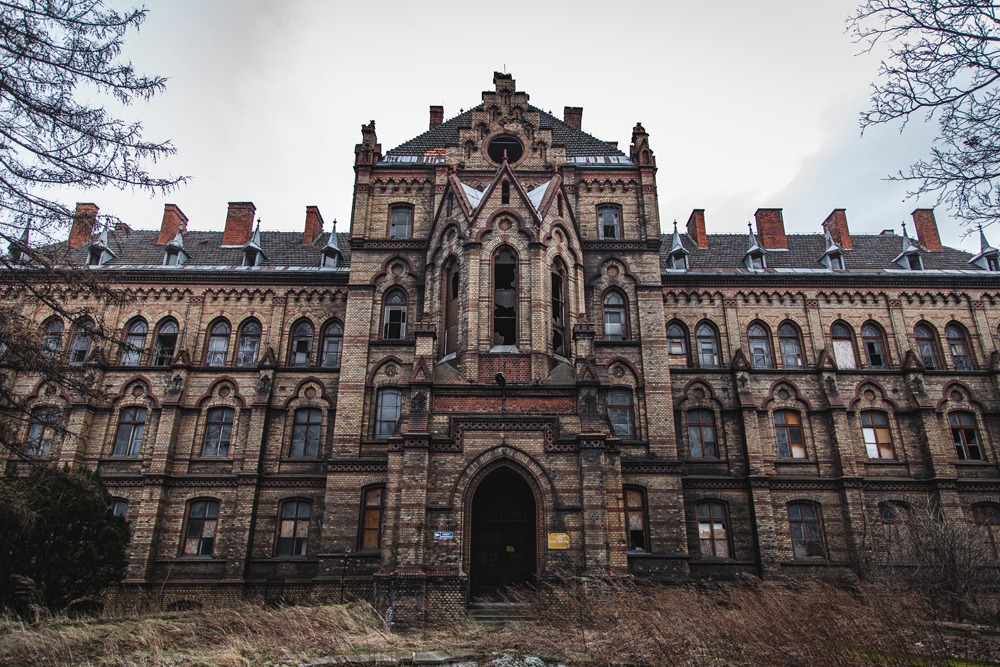 Fasada opuszczonego, ceglanego budynku w stylu neogotyckim z powybijanymi oknami i śladami zniszczeń, widoczna z dołu.