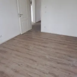 Jasne panele podłogowe ułożone w pustym, wykończonym pokoju z białymi ścianami i otwartymi drzwiami do kolejnych pomieszczeń; widoczne gniazdka elektryczne i wystający kabel z sufitu.