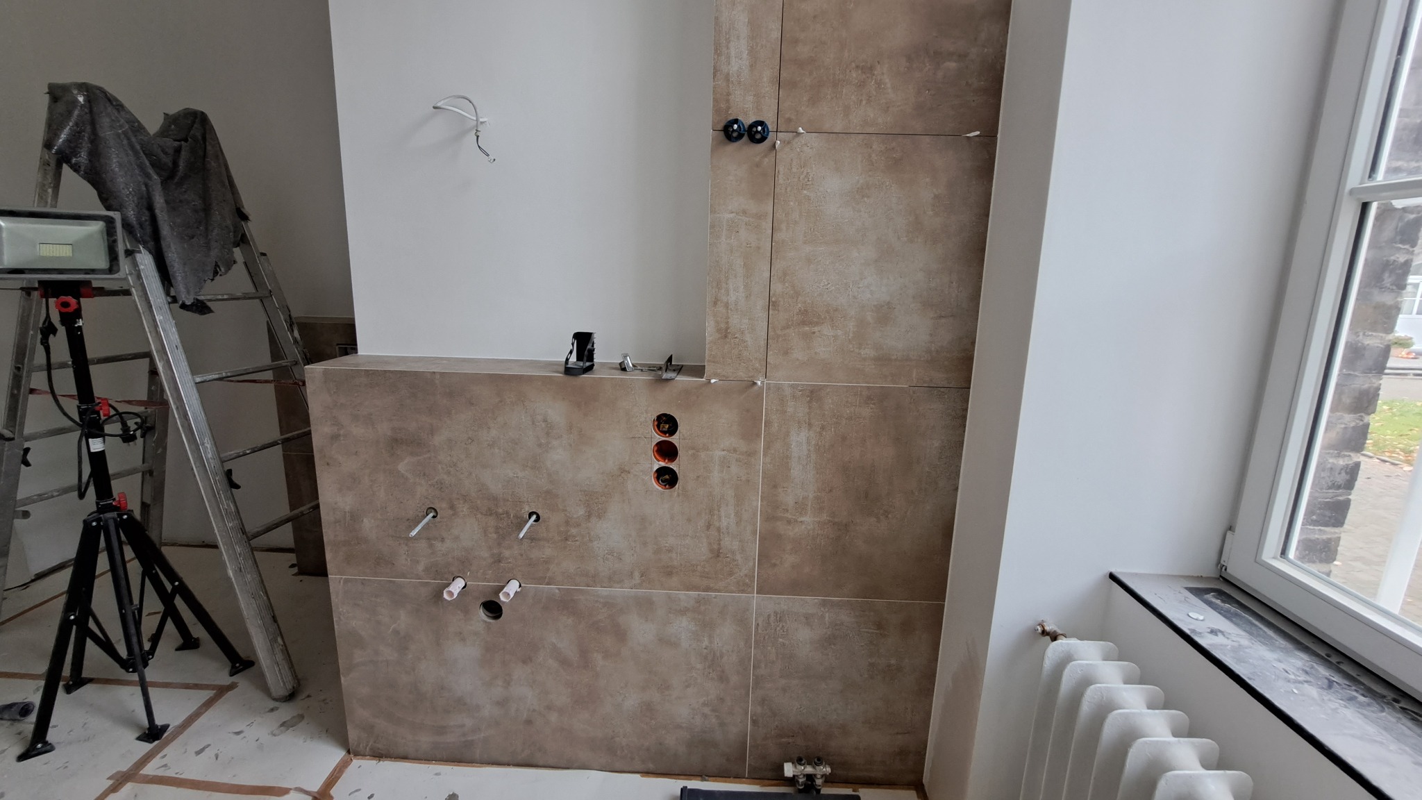 Ściana w trakcie kładzenia płytek ceramicznych w łazience, widoczne otwory na instalacje, drabina z reflektorem, krzyżyki dystansowe i poziomica.