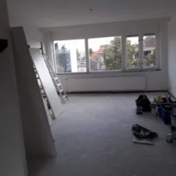 Pokój w trakcie malowania z widocznymi narzędziami malarskimi, drabiną i częściowo pomalowanymi ścianami na biało, przy oknie widoczne budynki mieszkalne.