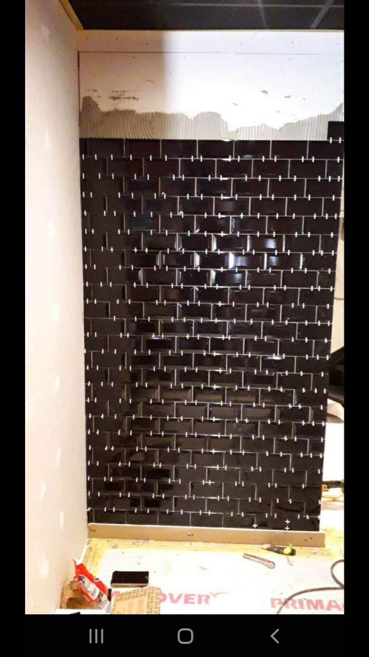 Układanie błyszczącej, czarnej glazury typu metro na ścianie z użyciem krzyżyków dystansowych, widoczna warstwa kleju na górze ściany, narzędzia glazurnicze leżące na podłodze.