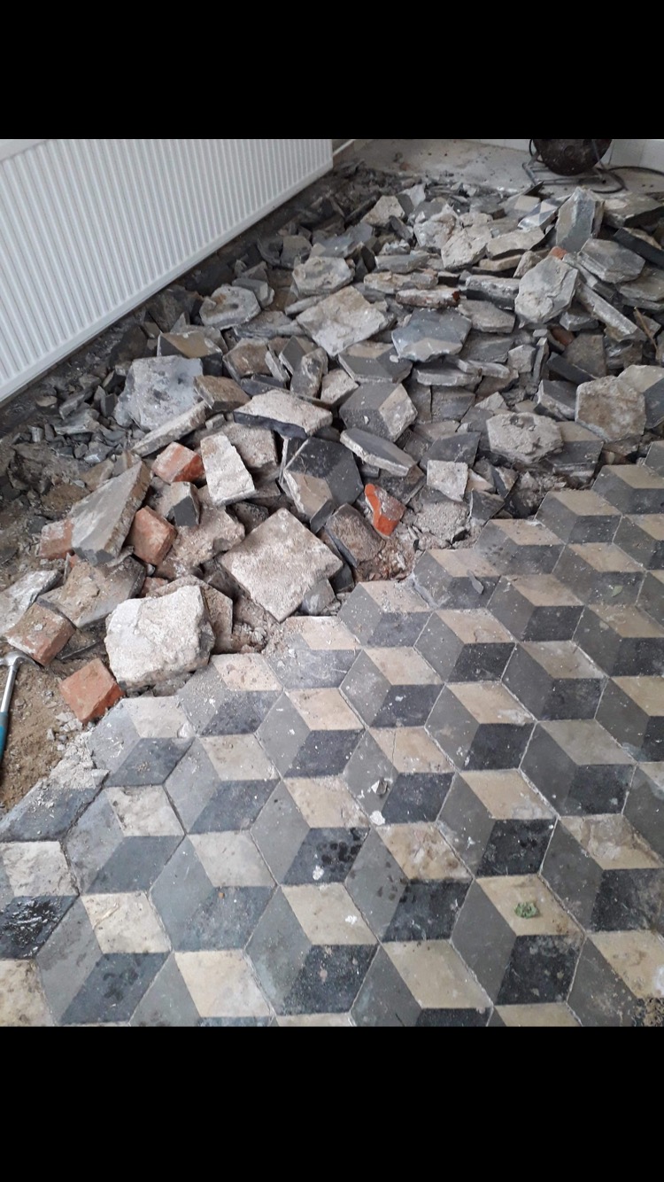 Demontaż podłogi z płytek w geometryczny wzór 3D, widoczne gruz i fragmenty starej posadzki, grzejnik w tle.