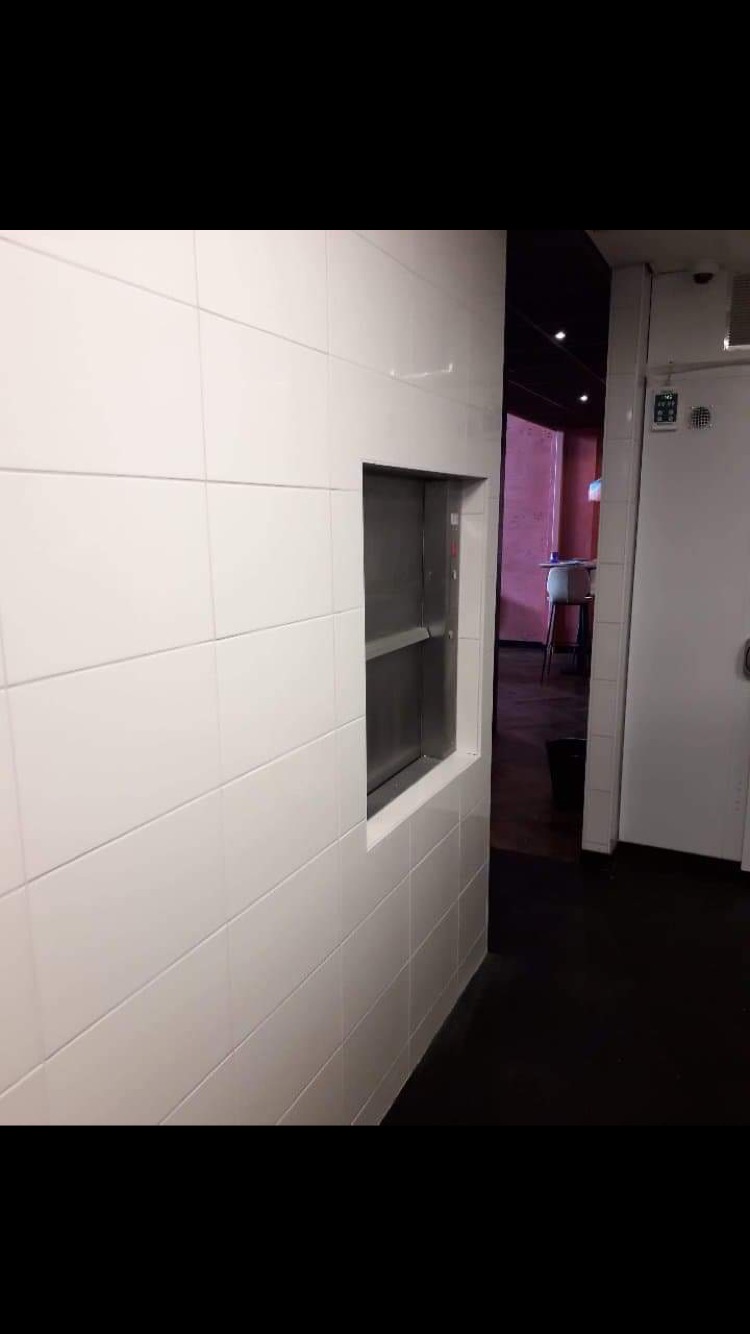 Białe, kwadratowe płytki na ścianie z wycięciem na okno podawcze, widoczny fragment pomieszczenia z różową ścianą i stołem w tle.