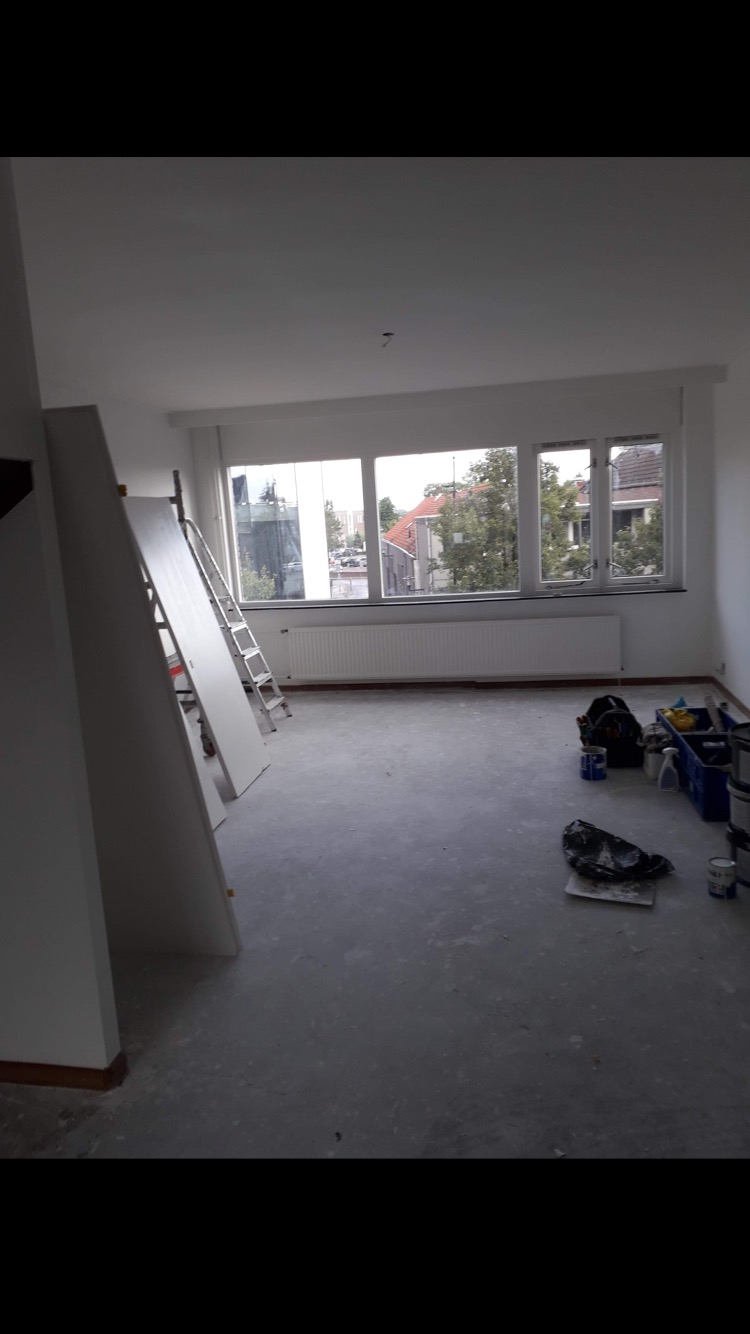 Pokój w trakcie malowania z widocznymi narzędziami malarskimi, drabiną i częściowo pomalowanymi ścianami na biało, przy oknie widoczne budynki mieszkalne.