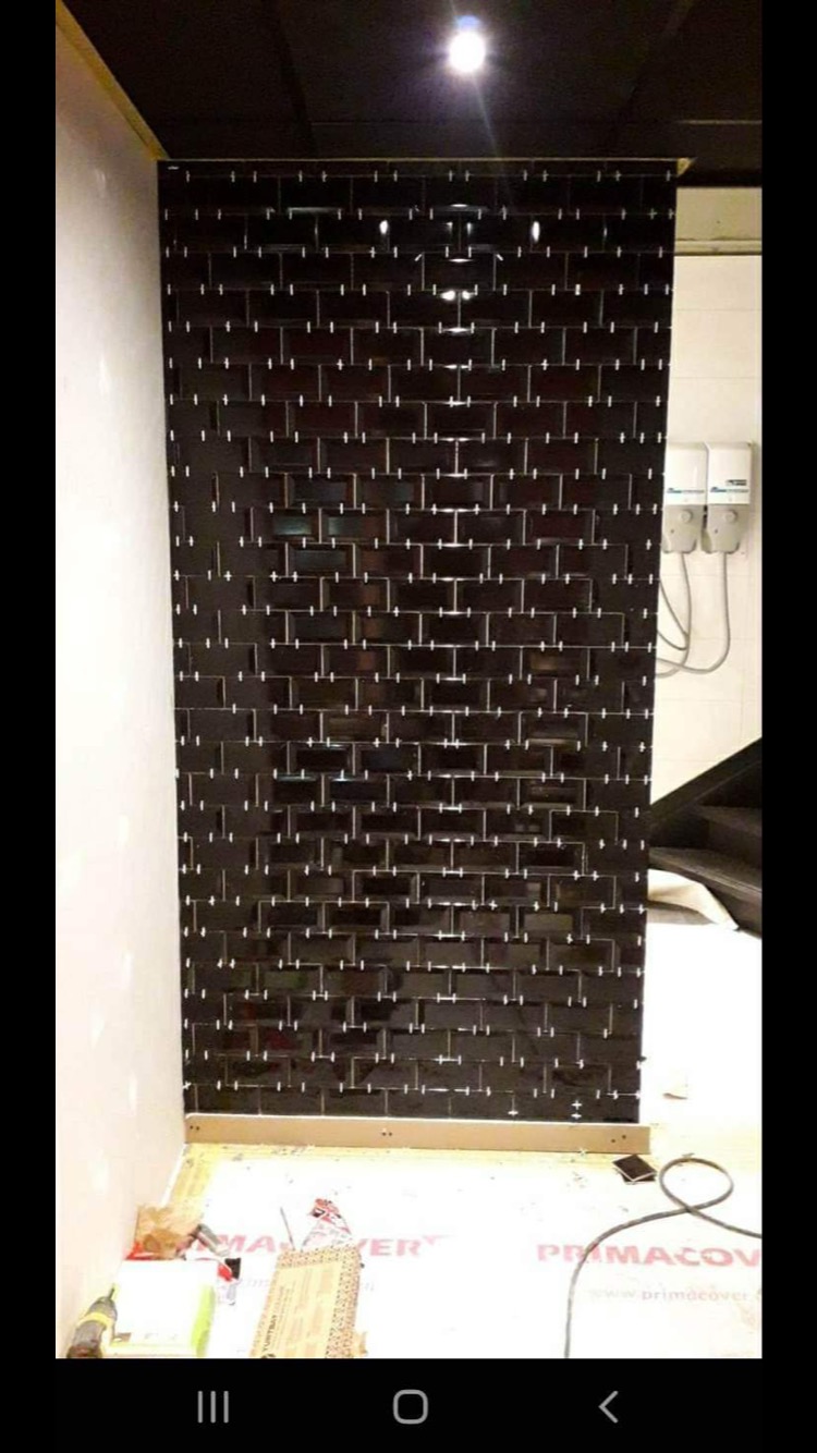 Ściana wyłożona błyszczącymi, czarnymi płytkami typu metro, z widocznymi białymi fugami, w trakcie prac wykończeniowych, widoczne narzędzia i materiały budowlane na podłodze.