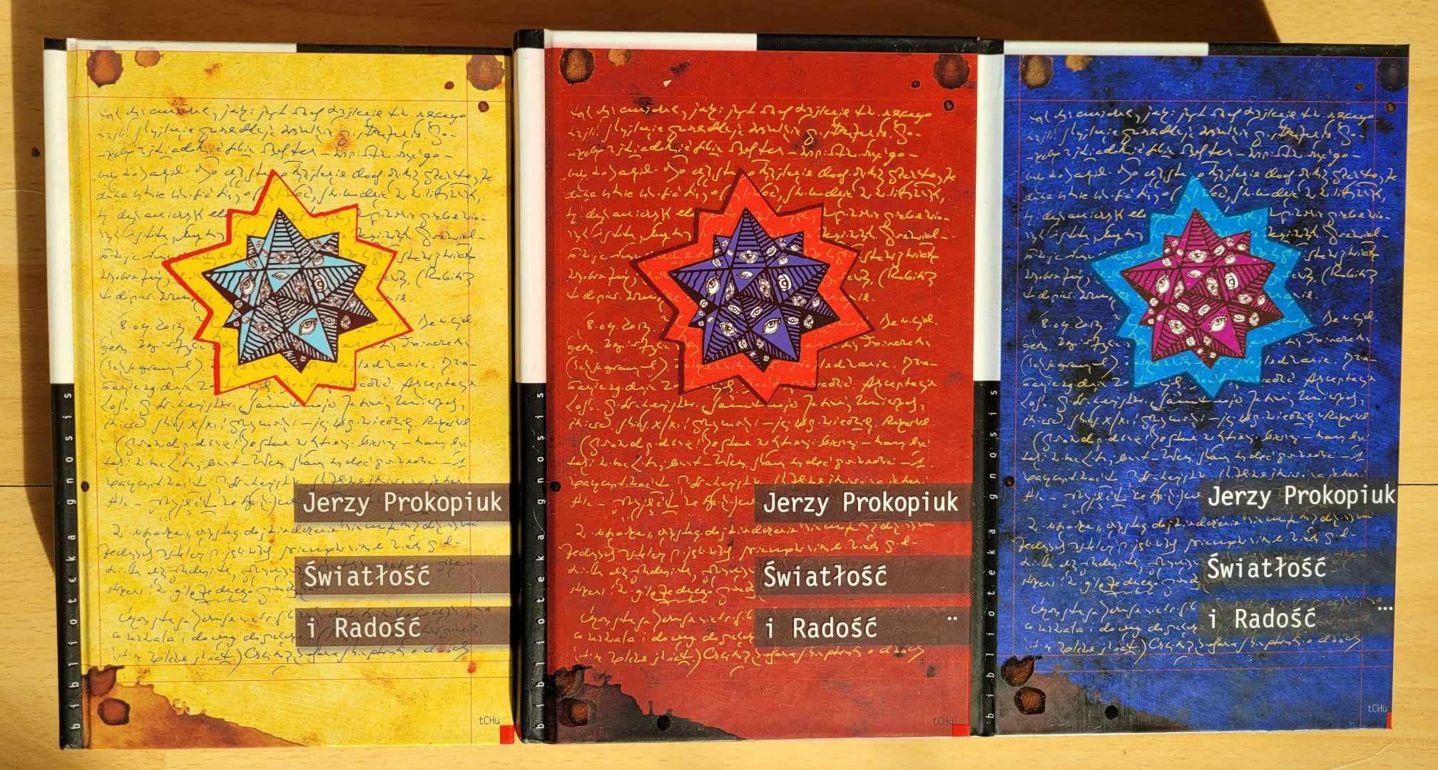 Trzy tomy książki 'Światłość i Radość' Jerzego Prokopiuka, każda w innym kolorze (żółty, czerwony, niebieski), z okładkami zdobionymi rękopisami i geometrycznymi gwiazdami z motywem oka.