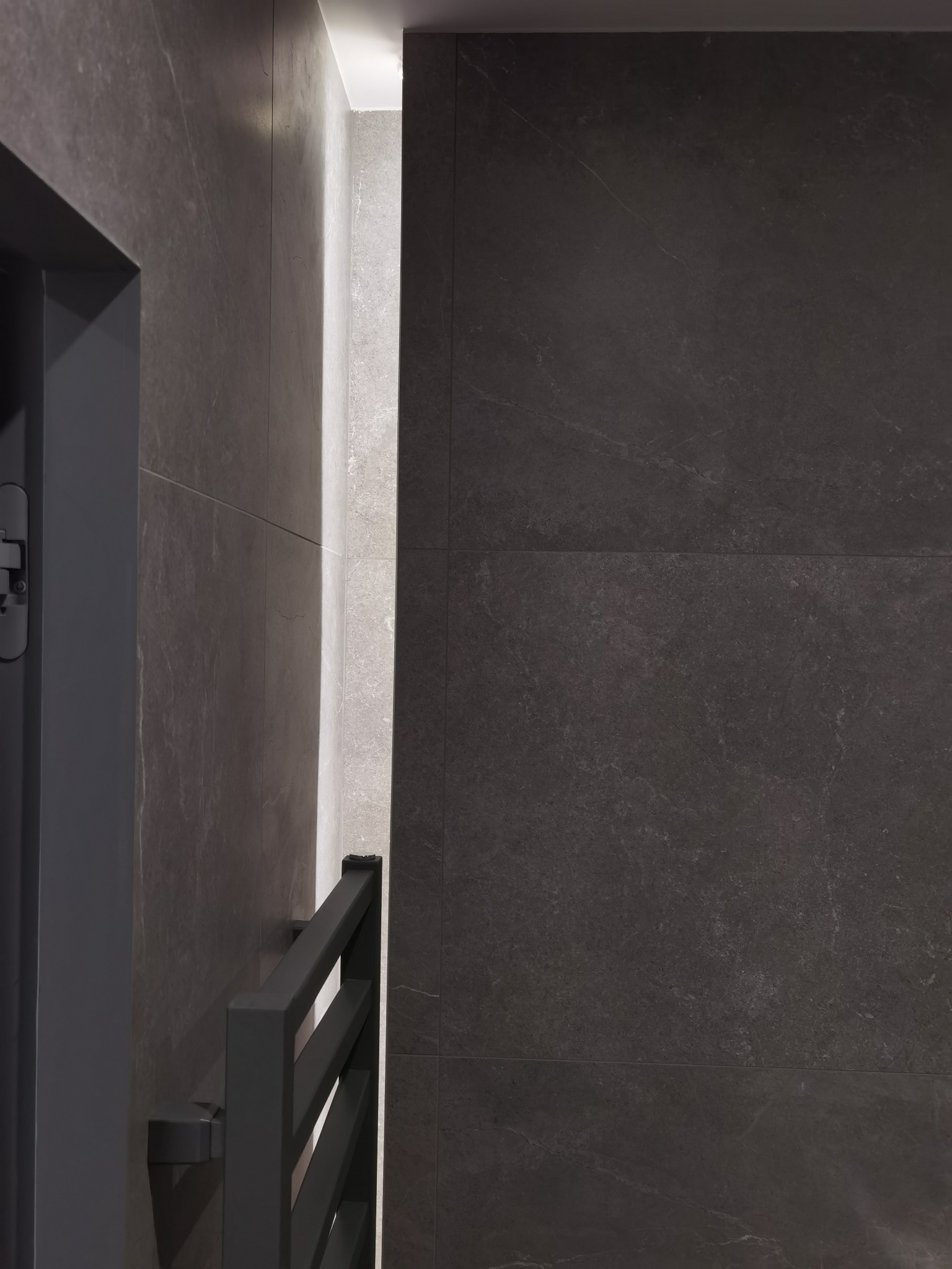 Ciemnoszara łazienka wykończona płytkami imitującymi kamień, widoczny fragment grzejnika drabinkowego w kolorze antracytowym i fragment oświetlenia LED w niszy.