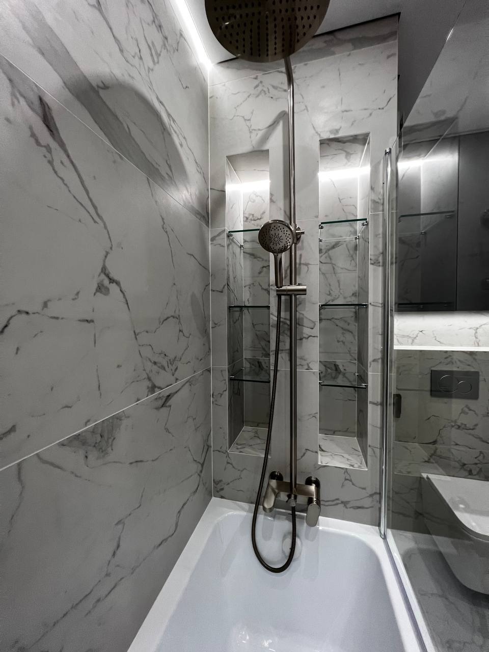 Nowoczesna łazienka z marmurowymi płytkami, kabiną prysznicową typu walk-in, baterią w kolorze szczotkowanego metalu i podświetlanymi niszami z szklanymi półkami.