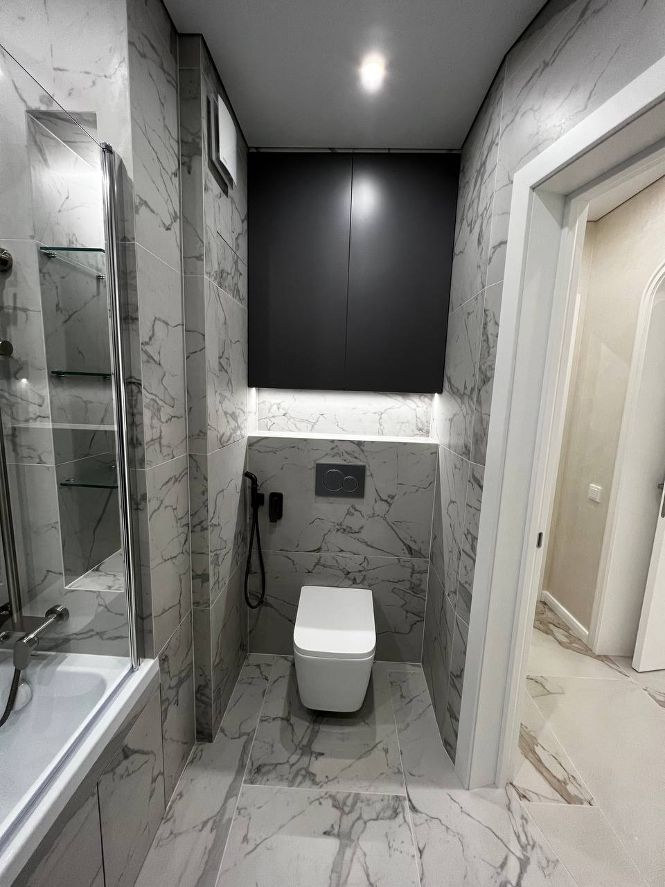 Nowoczesna łazienka z marmurowymi płytkami, wiszącą toaletą i czarną szafką nad nią, oświetlenie LED pod szafką, widok na fragment wanny z parawanem.