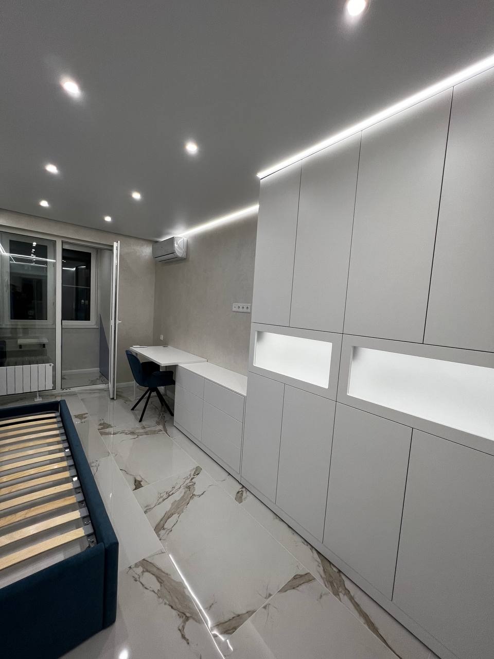 Nowoczesne wnętrze pokoju z białymi szafami w zabudowie z podświetleniem LED, biurkiem, krzesłem i łóżkiem bez materaca, marmurowa podłoga, widok na balkon.
