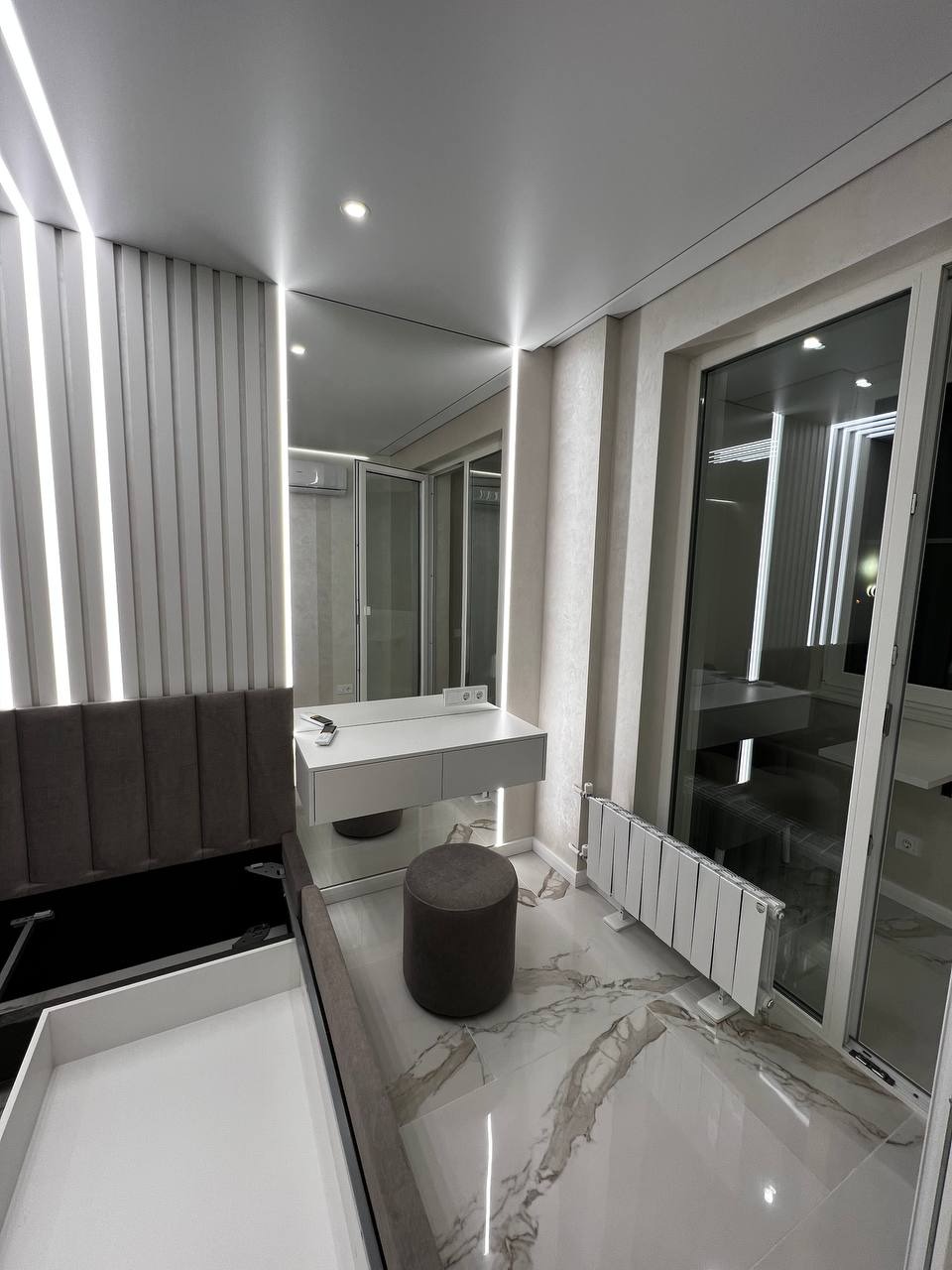 Nowoczesna sypialnia z toaletką, siedziskiem i dekoracyjną ścianą z pionowych paneli, oświetlona ledowymi lampami sufitowymi, z marmurową podłogą i dużym oknem balkonowym.
