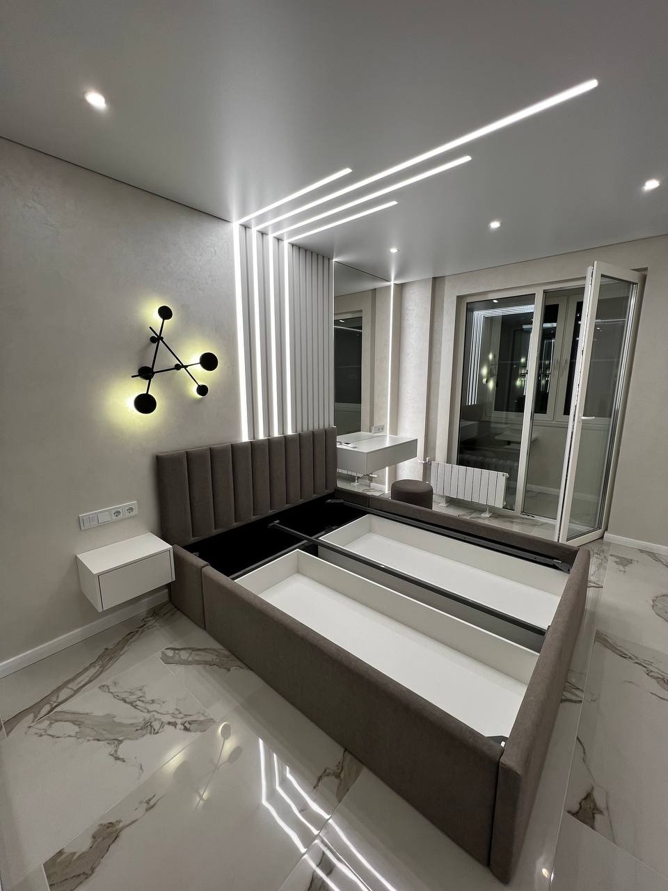 Sypialnia w nowoczesnym stylu: łóżko z szarym zagłówkiem i wysuwanymi białymi szufladami, marmurowa podłoga, designerska lampa ścienna i oświetlenie sufitowe LED.