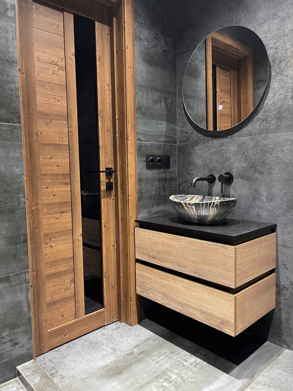 Nowoczesna łazienka z drewnianymi drzwiami z czarnym przeszkleniem, okrągłym lustrem, umywalką w czarno-złote wzory na szafce z dwoma szufladami i szarymi płytkami na ścianach i podłodze.