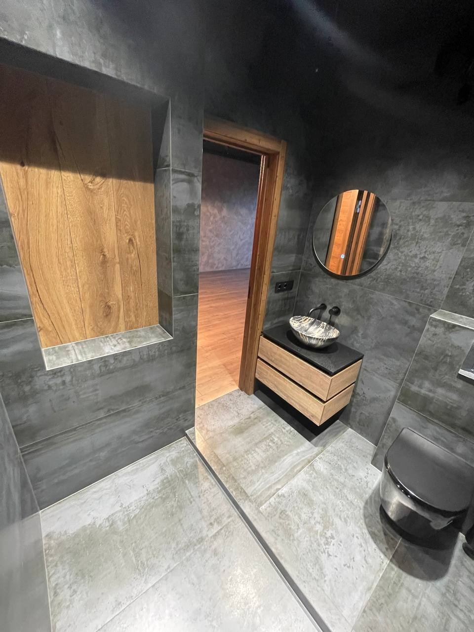 Nowoczesna łazienka z szarymi płytkami imitującymi beton, drewnianymi akcentami w postaci wnęki i szafki pod umywalkę oraz czarną armaturą i toaletą, widok na sąsiednie pomieszczenie z drewnianą...