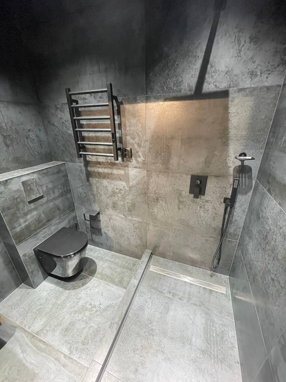 Nowoczesna łazienka z szarymi płytkami imitującymi beton, wisząca toaleta z chromowanym wykończeniem, czarny grzejnik drabinkowy i odpływ liniowy pod prysznicem bez brodzika.