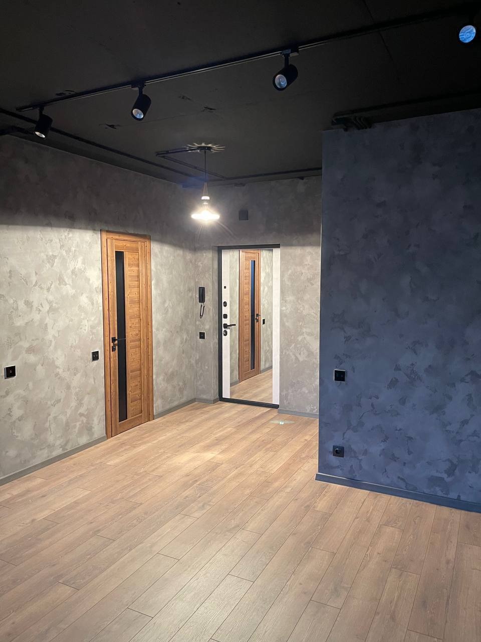 Wnętrze pokoju po remoncie z drewnianą podłogą panelową, ścianami w odcieniach szarości z efektem dekoracyjnym, czarnym sufitem z reflektorami i drewnianymi drzwiami z przeszkleniem.