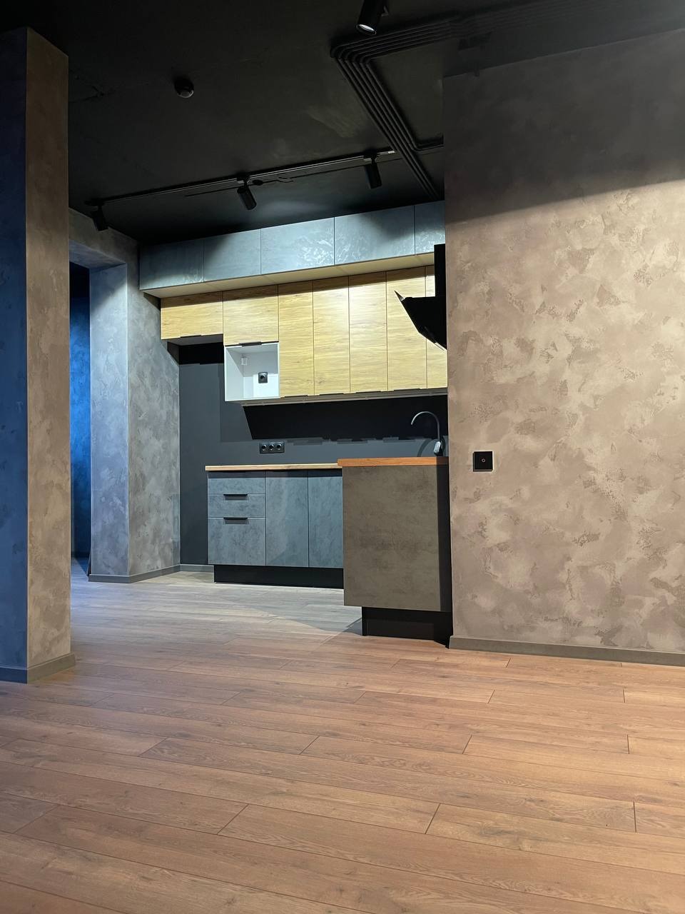 Nowoczesna kuchnia z drewnianymi panelami podłogowymi, szarymi szafkami i ścianami wykończonymi w dekoracyjnym tynku imitującym beton, oświetlona reflektorami na czarnym suficie.